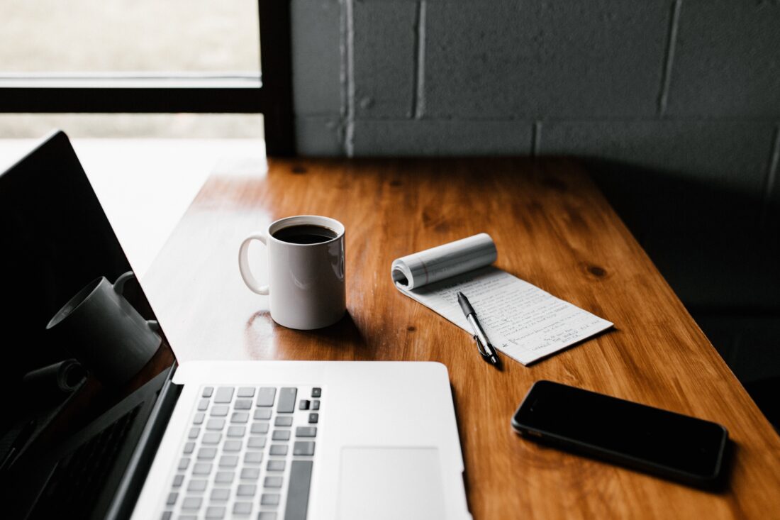Laptop sitting next to a cup of coffee, notepad, and cell phone.